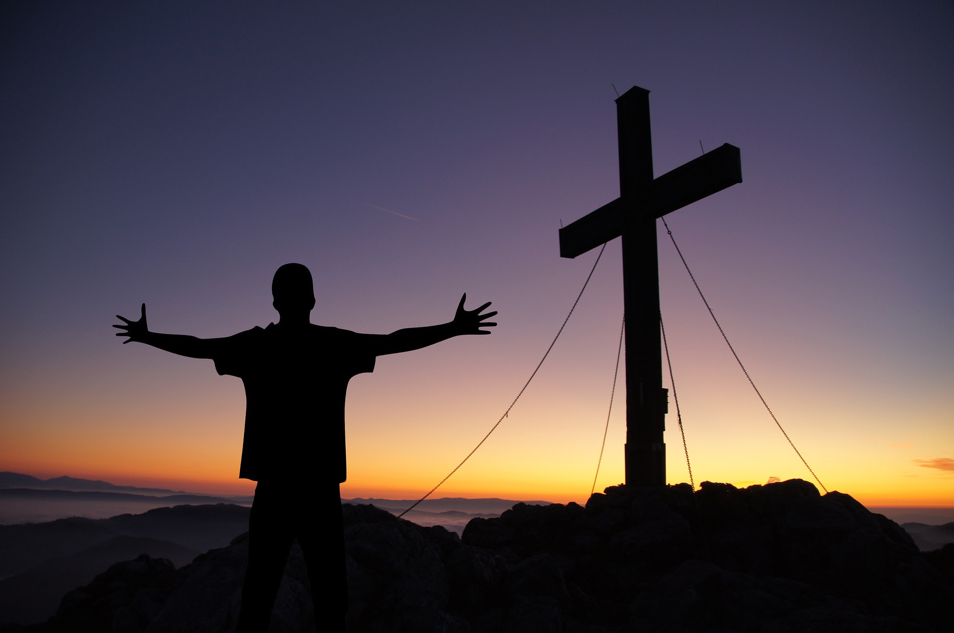 man with outstretched hands standing in front of a cross at sunset